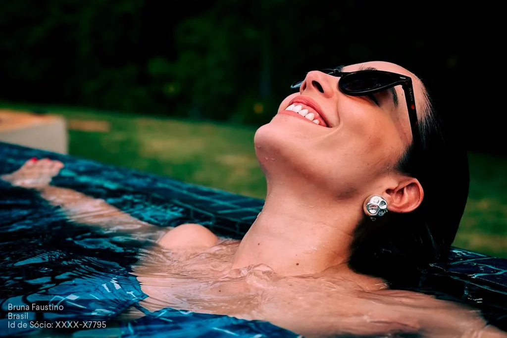 Mulher relaxando em uma piscina, sorrindo com óculos escuros, representando o bem-estar promovido pelo Sócio Influencer.